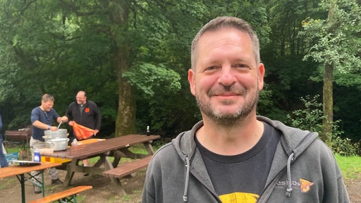 Ein Mann trägt einen Hoodie und steht vor einem Wald. Im Hintergrund wird Gulasch gekocht 