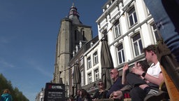 Ein Kirchturm ragt in den blauen Himmel. Darunter stehen altniederländische Herrenhäuser, an der Straße sitzen Menschen auf einer Terrasse.