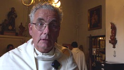 Ein alterer Mann trägt weiße liturgische Gewände und steht in einem dunken Zimmer mit Heiligenbildern und Kreuzen an der Wand.