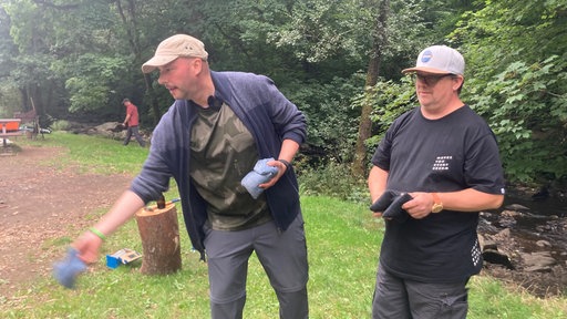 Ein Mann beugt sich nach vorne: Er ist im Begriff, einen Sack zu werfen. Ein anderer Mann steht daneben, ebenfalls mit zwei Säcken in der Hand, und guckt ihm zu.