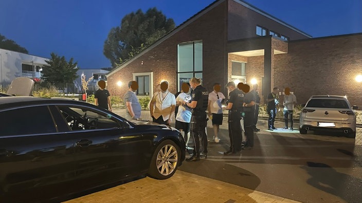 Parkplatz einer Wohnsiedlung bei Nacht. Polizisten kontrollieren eine Gruppe junger Leute an ihren Autos