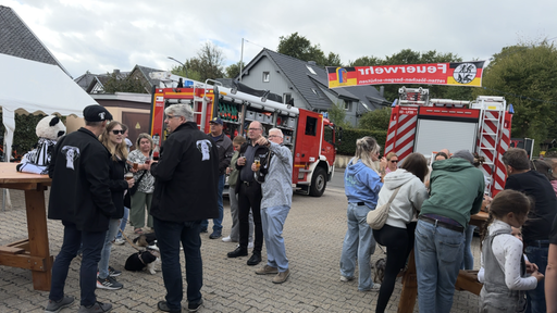 Menschen unterhalten sich auf dem Feuerwehrfest mit einem Feuerwehrwagen im Hintergrund