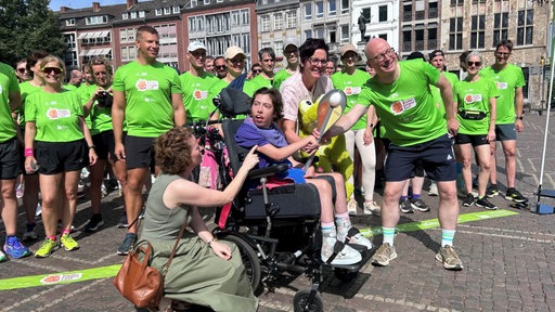 Für ein Foto posieren Läufer in grünen T-Shirts hinter eonem Mädchen, das in einem Rollstuhl sitzt. Links neben dem Rollstuhl hockt eine Frau, die lächelnd zum Mädchen schaut.