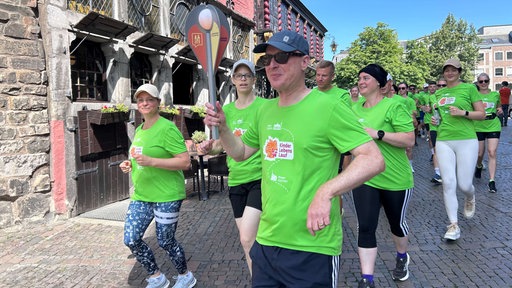 Mehrere Läufer in neongrünen T-Shirts laufen über den Marktplatz in Aachen. Der vorderste Läufer trägt eine unechte Fackel. 