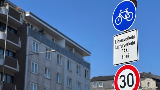 Am Aachener Löhergraben hängen drei Verkehrsschilder an einem Pfosten. Das Oberste zeigt auf blauem Hintergrund ein Fahrradsymbol. 