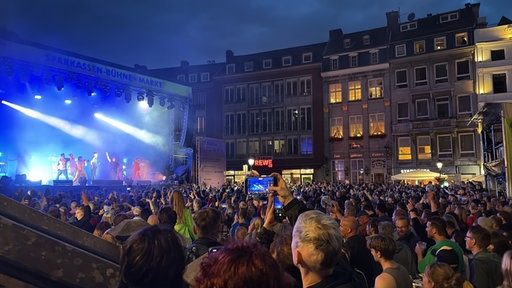Viele Menschen stehen auf einem Platz vor einer Bühne, auf der eine Band auftritt.