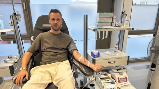 Auf einer aufgerichteten Liege liegt ein Mann mit kurzem Bart. Sein Arm liegt auf einer Armlehne. Auf dem Arm ist ein kleinen Schlauch festgeklebt, der zu einer Kanüle in seiner Armbeuge führt. 