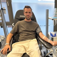 Auf einer aufgerichteten Liege liegt ein Mann mit kurzem Bart. Sein Arm liegt auf einer Armlehne. Auf dem Arm ist ein kleinen Schlauch festgeklebt, der zu einer Kanüle in seiner Armbeuge führt. 
