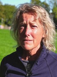 Eine Frau mit blonden Locken steht auf einer grünen Wiese und schaut an der Kamera vorbei