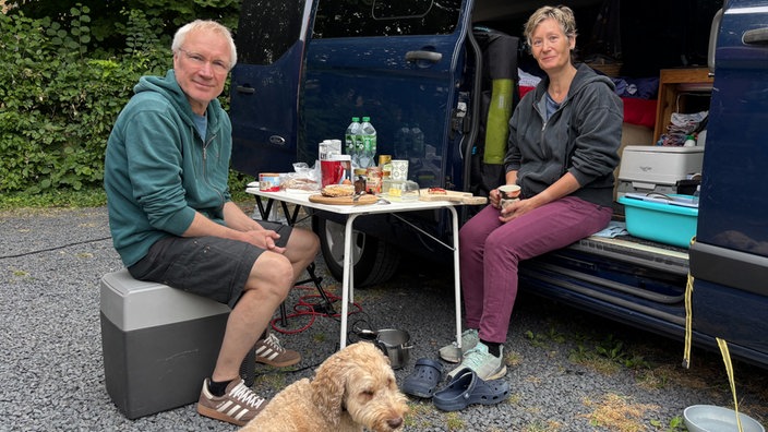 Paar sitzt neben offenem Auto am Campingtisch