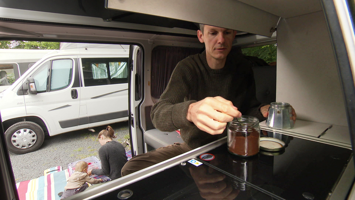 Ein Mann bereitet im Camper einen Kaffe vor, während man aus der Tür heraus eine Frau und ein Kleinkind sieht