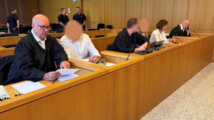 Gerichtssaal, die Angeklagten und ihre Anwälte, daneben die Dolmetscherin des einen Angeklagten