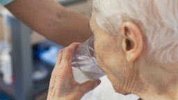 Eine ältere Frau mit kurzen weißen Haaren, trinkt aus einem Glas Wasser.