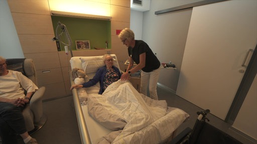 Eine Pflegerin deckt eine ältere Frau zu, die in einem Pflegebett liegt