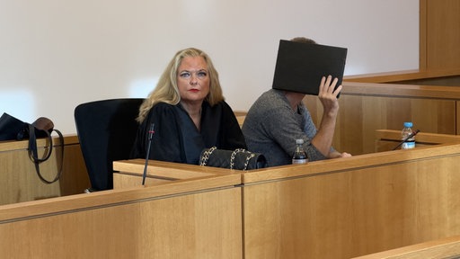 Der angeklagte Ex-Erzieher aus Aachen sitzt neben seiner Anwaltin im Gerichtssaal des Landgerichts und hält sich einen schwarzen Ordner vor das Gesicht