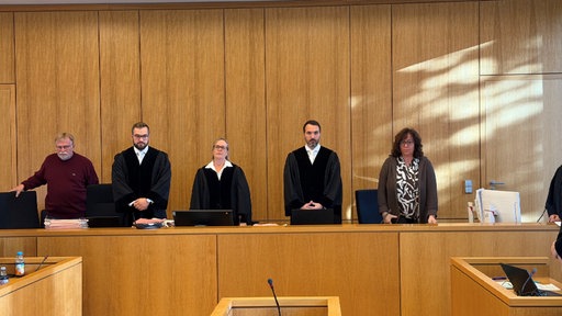 Das Gericht steht an ihren Plätzen; drei Menschen in Roben und jeweils rechts und links davon eine Person in normaler Kleidung