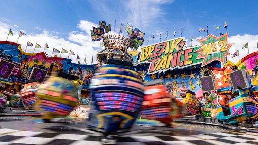 Die Gondeln eines Break-Dance-Karussells sind in voller Fahrt, im Hintergrund ist der blaue Himmel zu sehen.