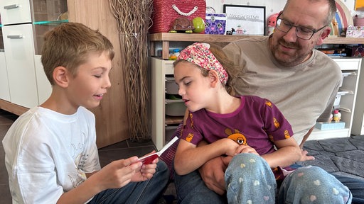 Ein Mädchen mit lila Kleid sitzt auf dem Schoß von einem Mann mit grauem Shirt. Links daneben sitzt ein Junge mit weißem Shirt und liest etwas vor. Im Hintergrund befindet sich ein Schrank mit diversen Gegenständen darauf.
