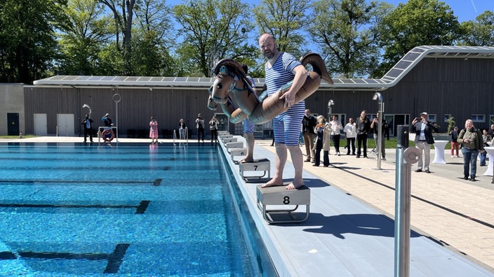 Ein Mann in einem blau-weiß gestreiften Schwimmanzug posiert mit einem Schwimmreifen vor einem Freibadbecken.