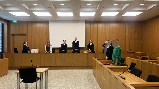 Im Gerichtssaal steht der Angeklagte in grüner Kleidung neben seiner Anwältin vor der Richterbank.