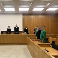 Im Gerichtssaal steht der Angeklagte in grüner Kleidung neben seiner Anwältin vor der Richterbank.