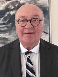 Anwalt Manfred Schmitz trägt eine schwarze Brille und steht vor einem braublaubraunen Gemälde 