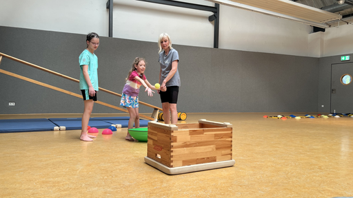 Kinder, die versuchen einen Ball in einen umgedrehten Kasten werfen. Neben ihnen die Kursleiterin.