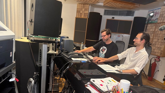 Der Komponist Marc Vogler sitzt in einem Tonstudio, im Hintergrund hilft ihm ein Produzent am Computer