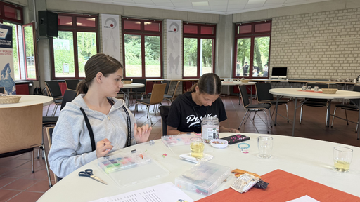 Lenuta und Andreea sitzen nebeneinander und bastelt an bunten Perlenarmbändern.