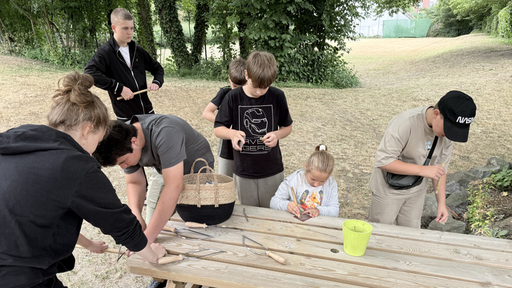 Kinder sägen und schleifen runde Holzstäbe. Sie befinden sich draußen an einem Holztisch.