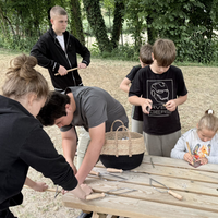 Kinder sägen und schleifen runde Holzstäbe. Sie befinden sich draußen an einem Holztisch.