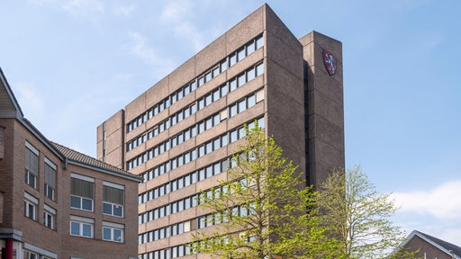 Ein braunes Gebäude mit vielen Fenstern. Auf der rechten Seite ist ein rotes Wappen an der Fassade.