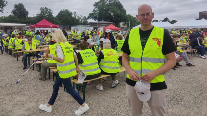 Tim Lösch von der Gewerkschaft Nahrung-Genuss-Gaststätten steht, mit einer gelben Warnweste bekleidet, auf einem Platz und schaut zur Kamera. Im Hintergrund sitzen viele Menschen auf Bierbänken und tragen gelbe Warnwesten.
