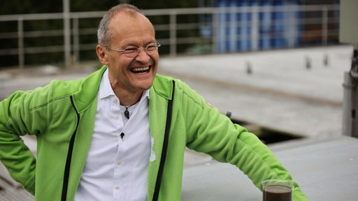 Klaus Vossenkaul lacht in weißem Hemd und grüner Strickjacke in die Kamera