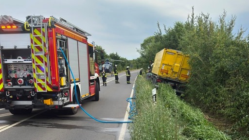 Landstraße: Ein Traktor und sein Anhänger im Straßengraben. Auf der Straße sind mehrere Feuerwehrleute, vorne links im Bild ein Feuerwehrauto