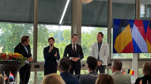 Sander Kleikers, Silke Gorißen, Jaimi van Essen und Jo Brouns in einer Podiumsdiskussion. Daneben eine belgische und einer niederländische Flagge.