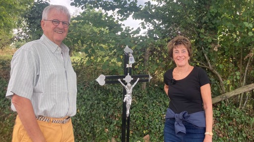 Man sieht ein Kreuz am Feldweg. Daneben stehen eine Frau und ein Mann, beide in Rente, mit einem Lächeln.