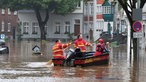 Ein rot-gelbes Motorboot fährt über einen mit Wasser überfluteten Platz, in dem Boot sitzen drei Menschen, zwei haben ein Ruder in der Hand, im Wasser schwimmen schwarze Mülltonnen