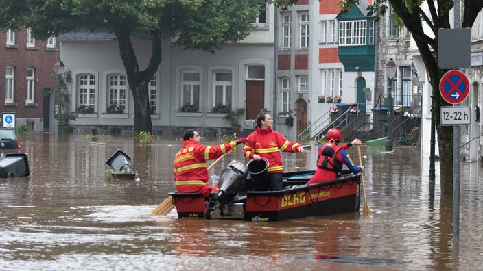 Ein rot-gelbes Motorboot fährt über einen mit Wasser überfluteten Platz, in dem Boot sitzen drei Menschen, zwei haben ein Ruder in der Hand, im Wasser schwimmen schwarze Mülltonnen