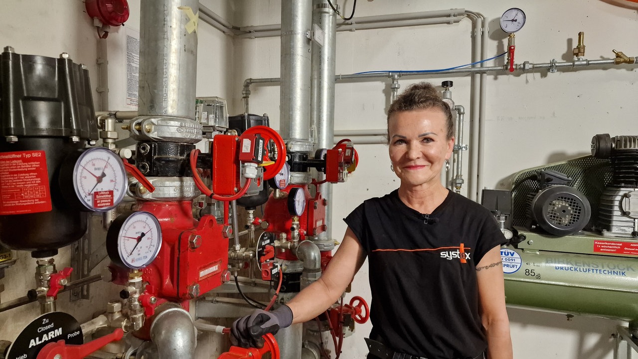 Eine Handwerkerin steht vor einer Anlage mit Rohren und Druckanzeigen.