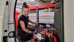 Eine Handwerkerin kniet in einem Auto, vor ihr ist Werkezeug in einem Eimer.