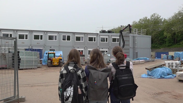 Drei Schülerinnen stehen vor einer Baustelle mit vielen Containern