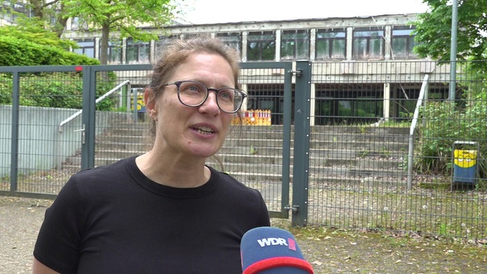 Ein Frau mit großer schwarzer Brille und schwarzem Oberteil war dem Schultor zur Brandruine der Schule