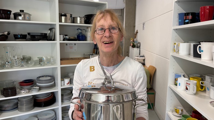Eine Frau hält einen großen, silbernen Topf in en Hänedn und lacht in die Kamera