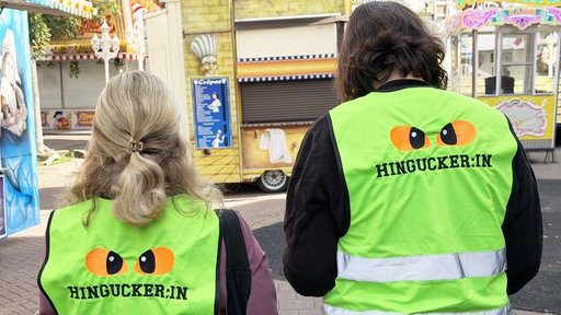 Zwei Personen stehen mit dem Rücken zur Kamera, beide tragen eine neongrüne Warnweste, mit der Aufschrift "Hingucker:in", im Hintergrund Kirmesbuden.