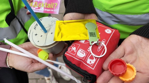 Zwei Personen halten verschiedene Utensilien, zur Vorbeugung der KO-Tropfen Zugabe, in den Händen.