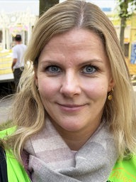 Eine Frau mit langen blonden Haaren, bekleidet mit einer neongrünen Warnweste, im Hintergrund stehen Kirmesbuden.