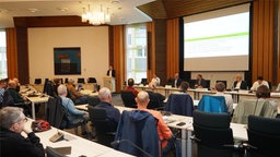 Menschen an Tischen, die auf ein Podium ausgerichtet sind, wo ein Mann eine Rede hält