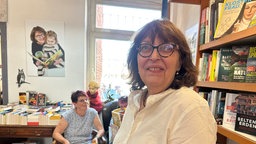 Frau mit offenen Haaren und Brille steht im Vordergrund und schaut in die Kamera, im Hintergrund sitzt eine weitere Frau und schaut zur Seite