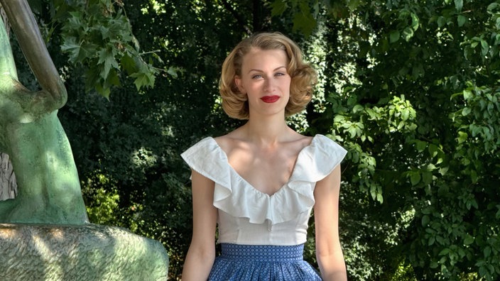 Lenina sitzt mit weißer Bluse und blauem Rock auf einer Mauer